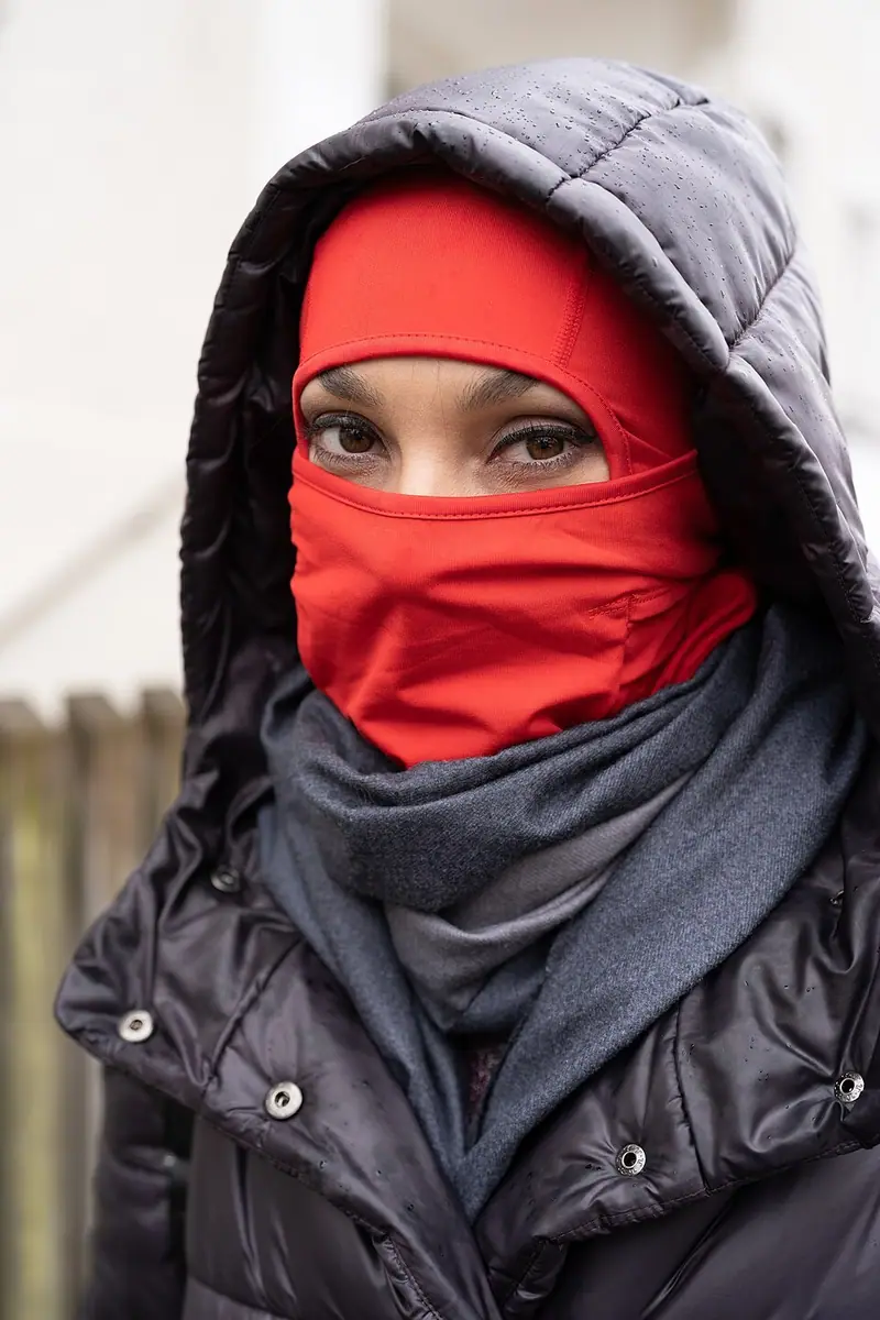 Une personne porte une cagoule rouge sous une capuche noire, le visage couvert à l’exception des yeux.