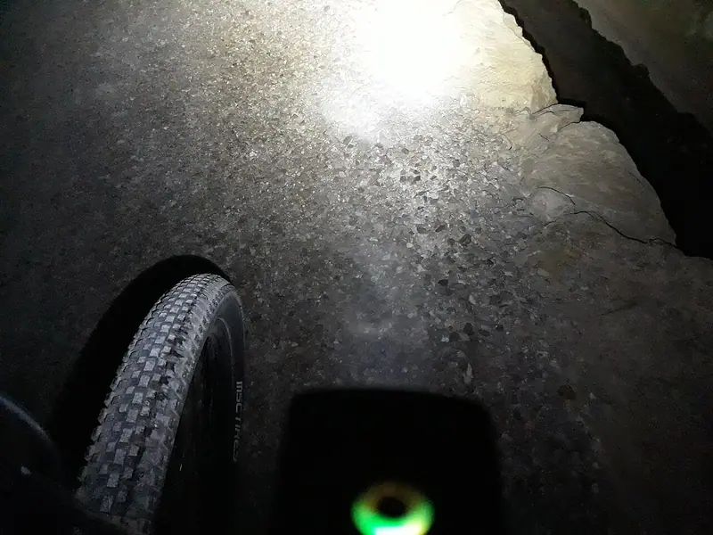 Faisceau blanc d’un éclairage de vélo sur un chemin sombre, avec la roue avant au premier plan.